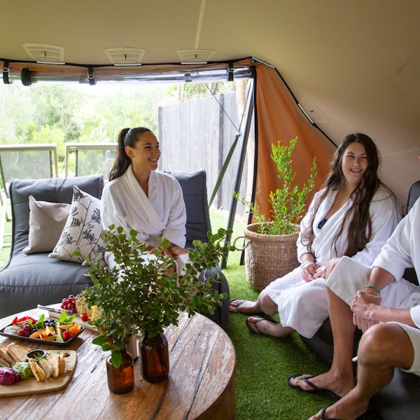 two women and a man in white bath robes sitting in tent style structure on comfortable day beds with fresh platters of food laid out on wooden coffee table