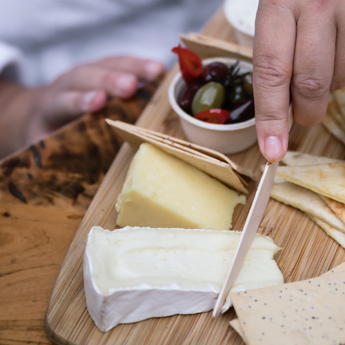 person in white bath robe cutting cheese on grazing board with olives and dip & pita breads