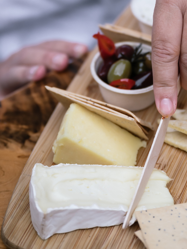 person in white bath robe cutting cheese on grazing board with olives and dip & pita breads