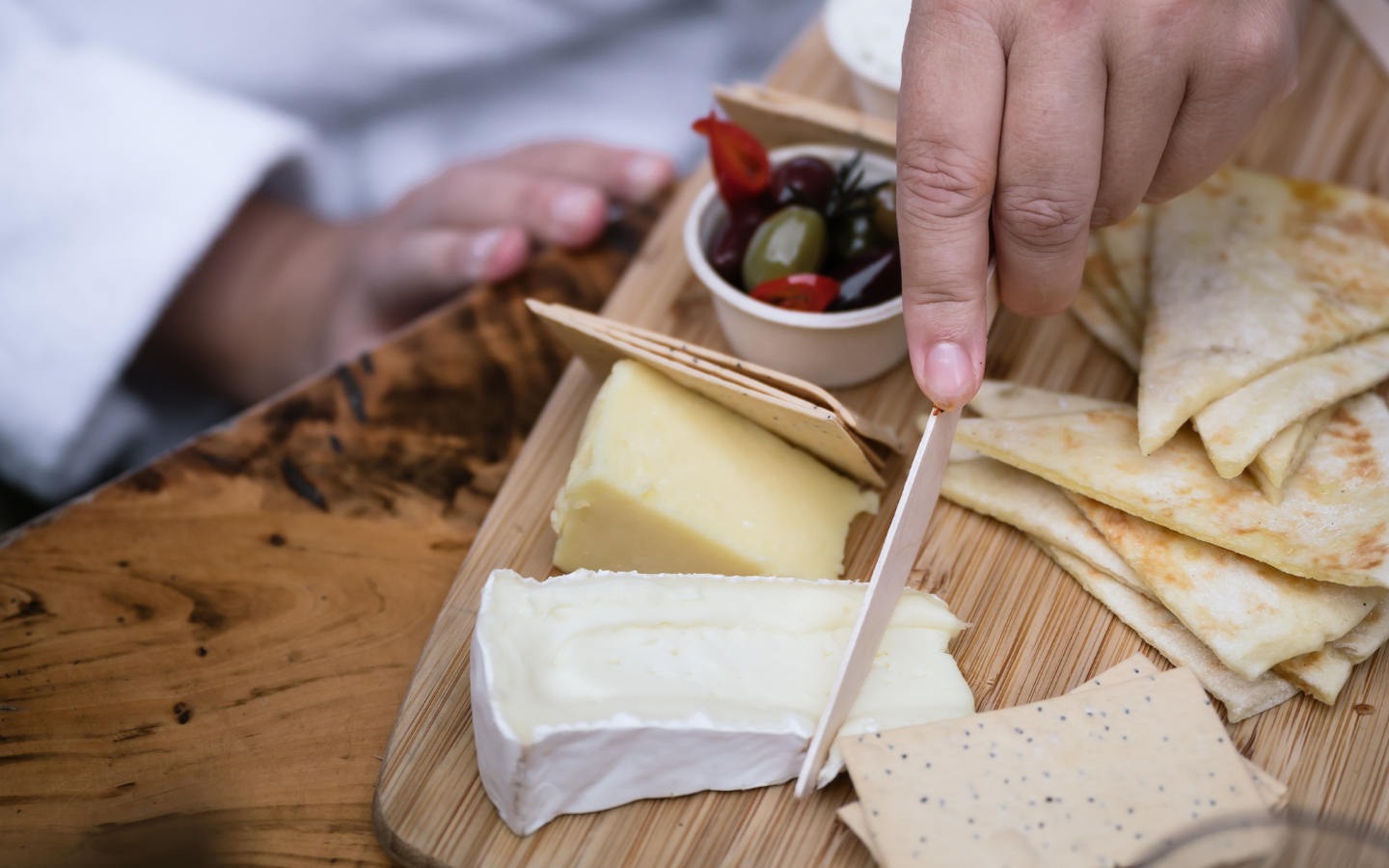 person in white bath robe cutting cheese on grazing board with olives and dip & pita breads