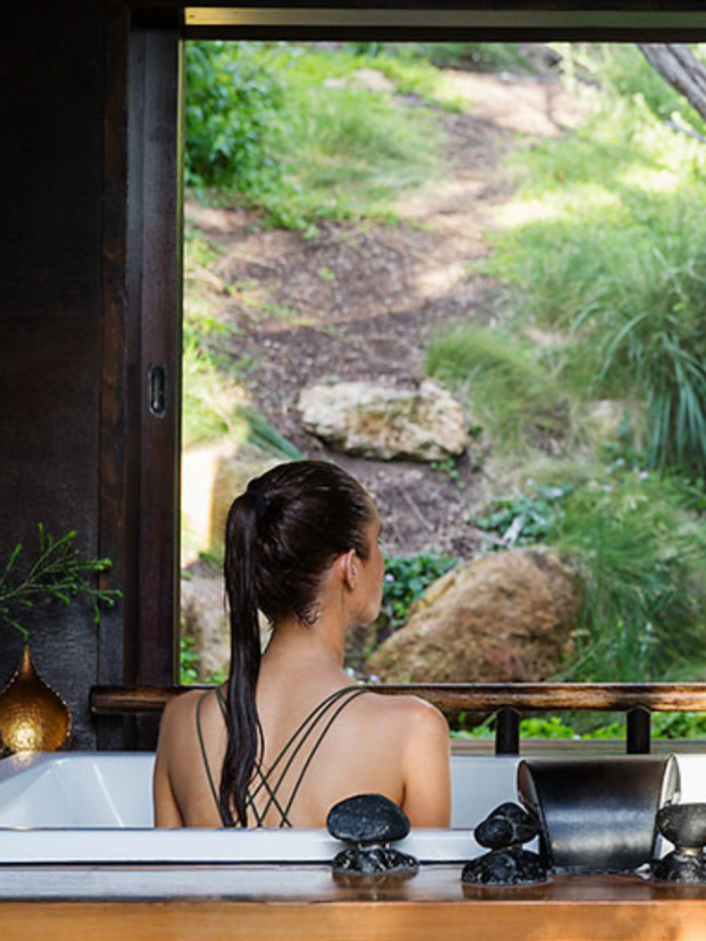 view of back of woman with long ponytail in bathing suit looking out at nature from her private bath