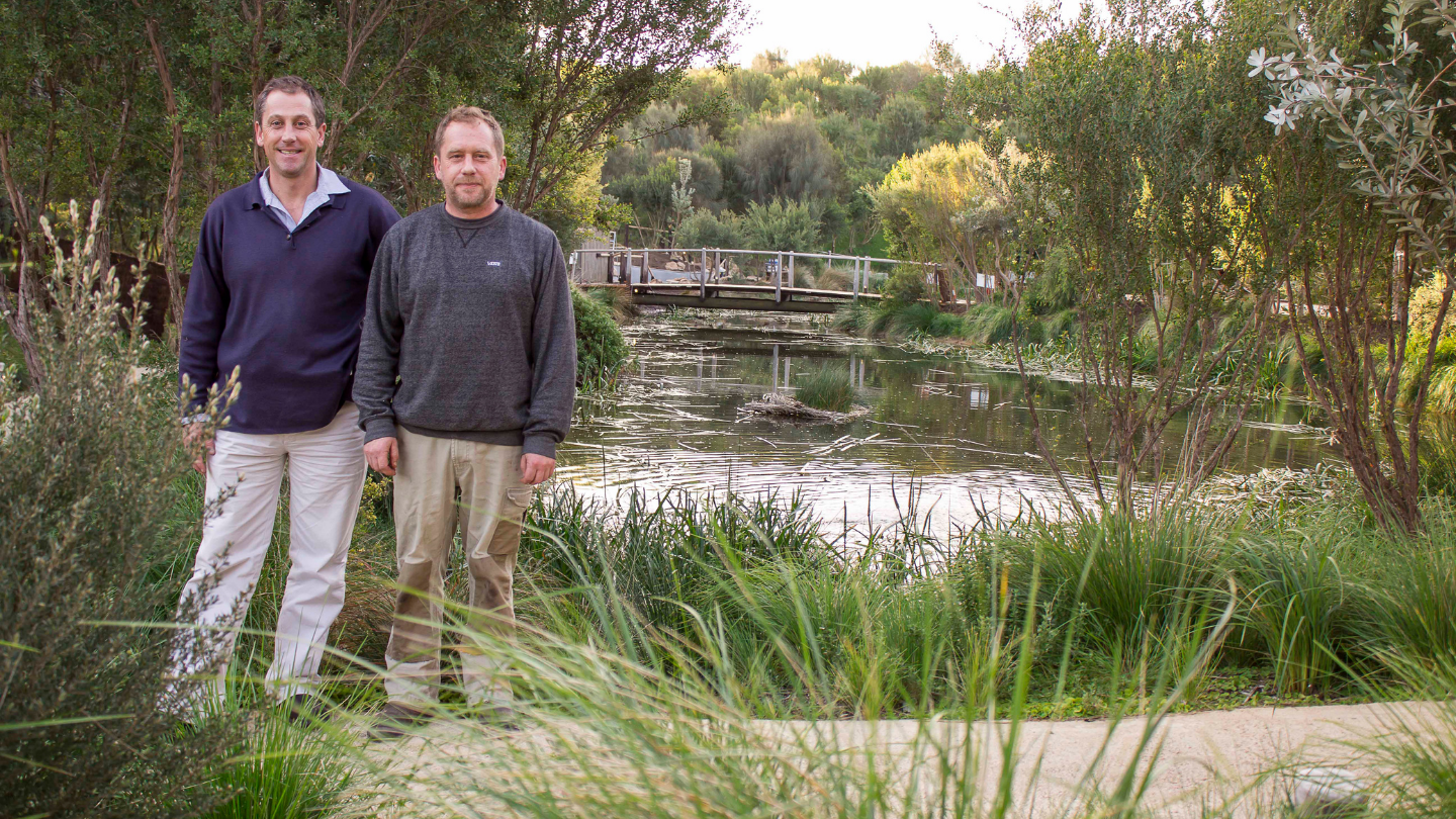 two men standing amongst bushland and in front of a lake