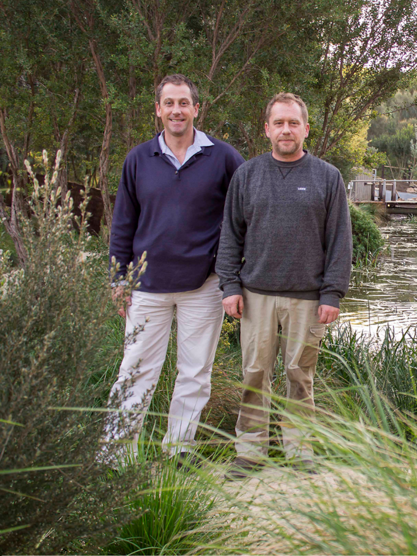 two men standing amongst bushland and in front of a lake