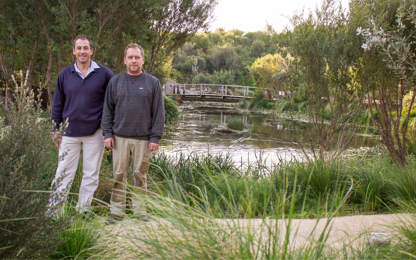 two men standing amongst bushland and in front of a lake