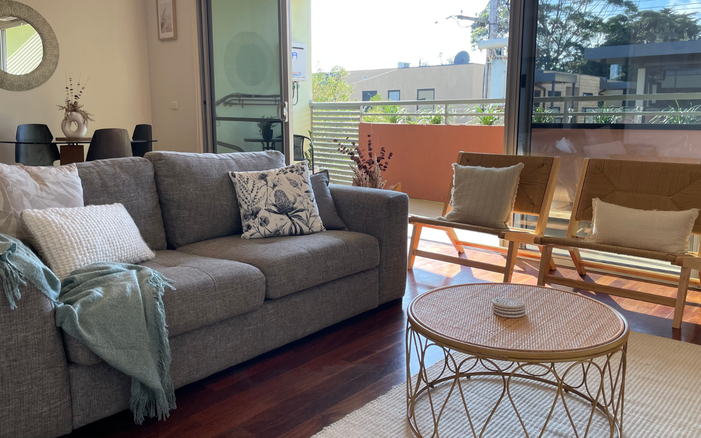 a living room set up with grey lounge chair, rattan chairs and open doors leading to a balcony flooded with natural light