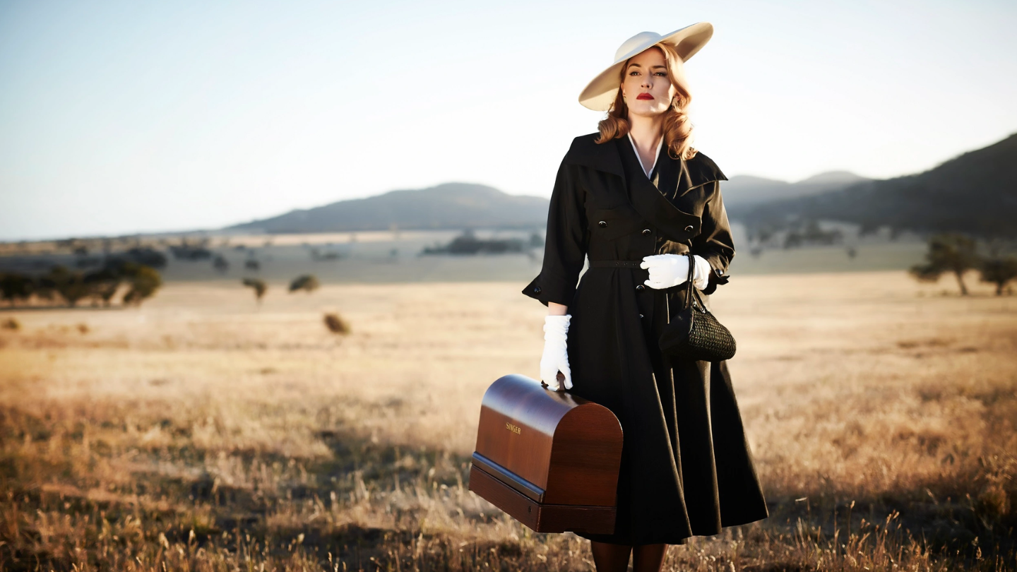 a women in a black trench coat, hat and white gloves carrying a briefcase and looking into the distance surrounded by the Australian outback