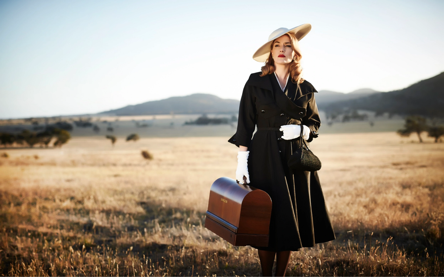 a women in a black trench coat, hat and white gloves carrying a briefcase and looking into the distance surrounded by the Australian outback