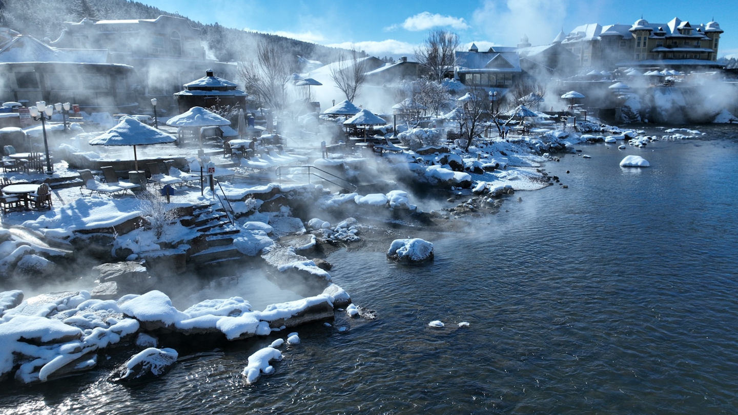 a wide shot of steaming hot springs surrounded by snow capped outdoor furniture, mountains and buildings