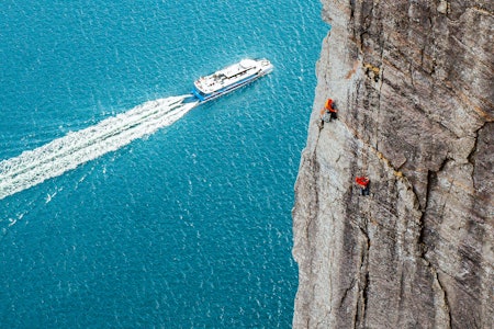 SPEKTAKULÆRT: Bratt og krevende klatring midt i en av Norges kanskje mest besøkte turistattraksjoner (og stedet hvor Tom Cruise har slåss med en superskurk hengende i en wire med et helikopter). Foto: Martin Lindgren klatrere i stup med fjord og båt i bakgrunnen