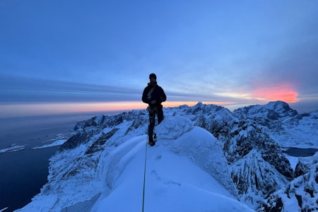 Stian Bruvoll på toppen av Klokktinden etter å ha forsert nordveggen sammen med Chris Wright.  Foto: Chris Wright Mann på toppen av nordnorsk snødekt fjell med dagens siste røde solstråler i bakgrunnen.