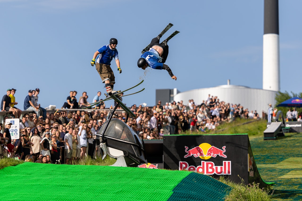 I jibbekonkurransen kjører deltagerne fra hvert land samtidig ned bakken. Foto: Jesper Gronnemark. En mann henger opp ned i luften i et hopp, mens en annen sklir langs toppen av en rail i sommerværet.