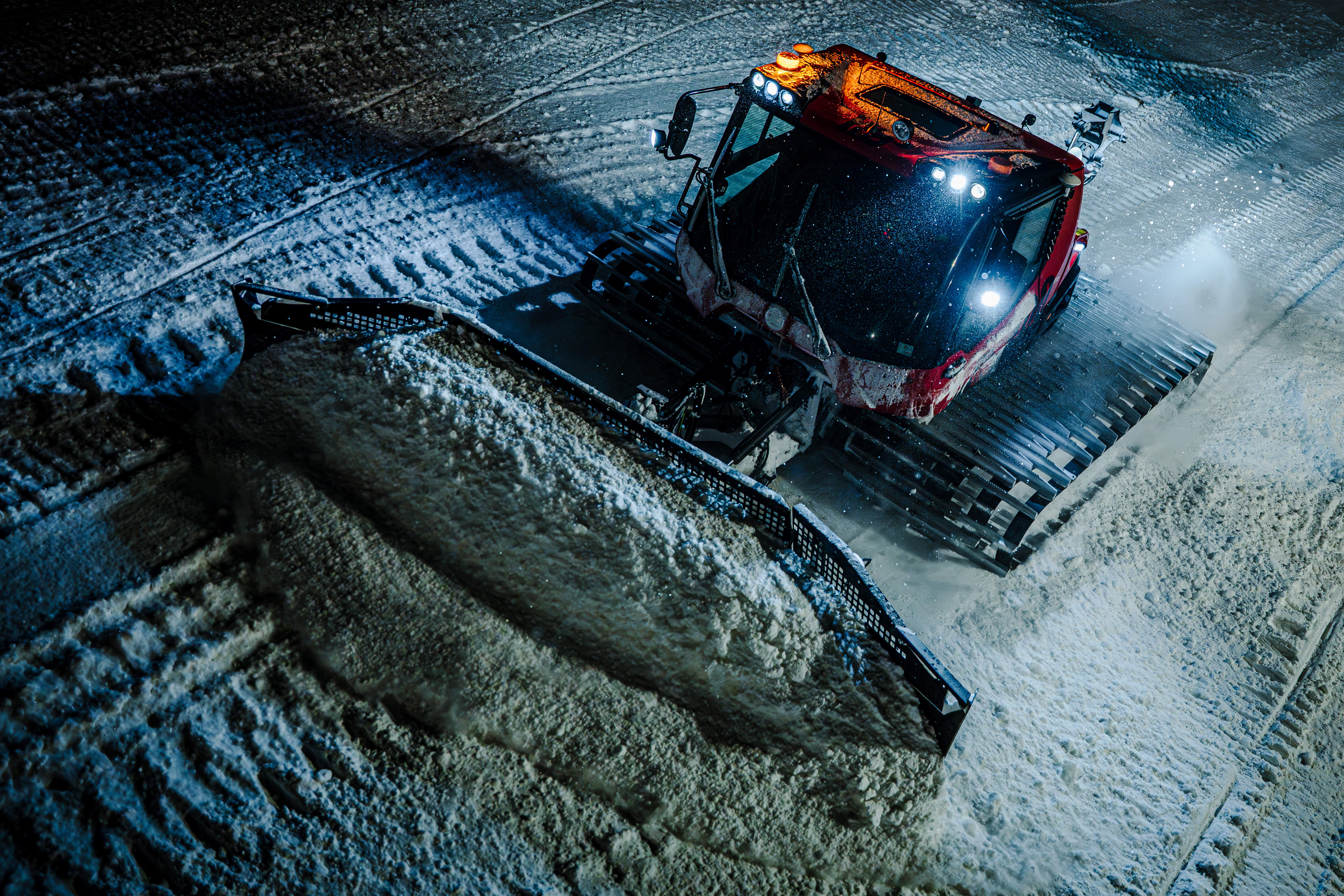 En stor tråkkemaskin kjører i en alpinbakke etter stengetid. Maskinen har sterke lys og flytter snø mens bakken prepareres i mørket