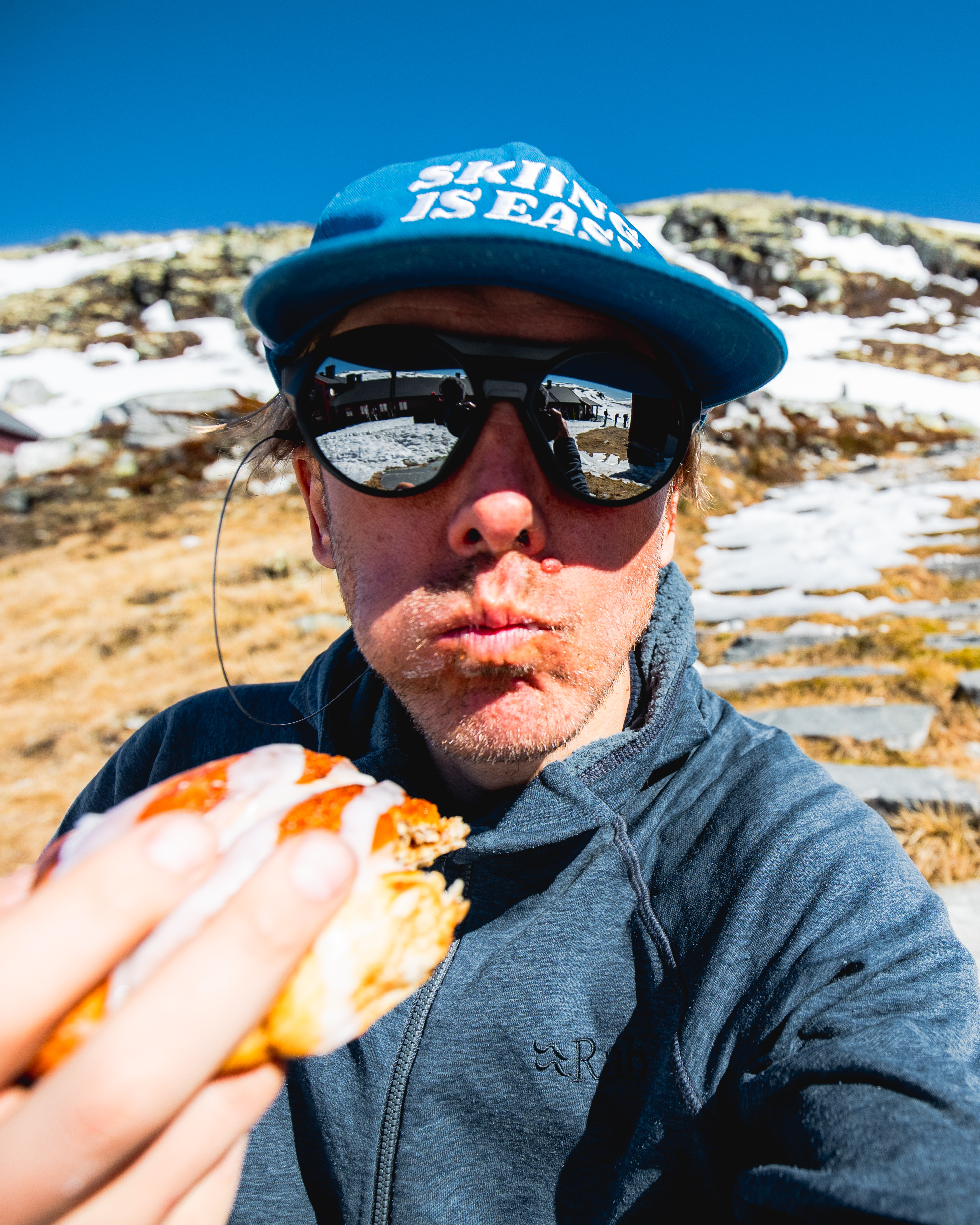 En person med caps og solbriller som spiser en bolle i fjellet.