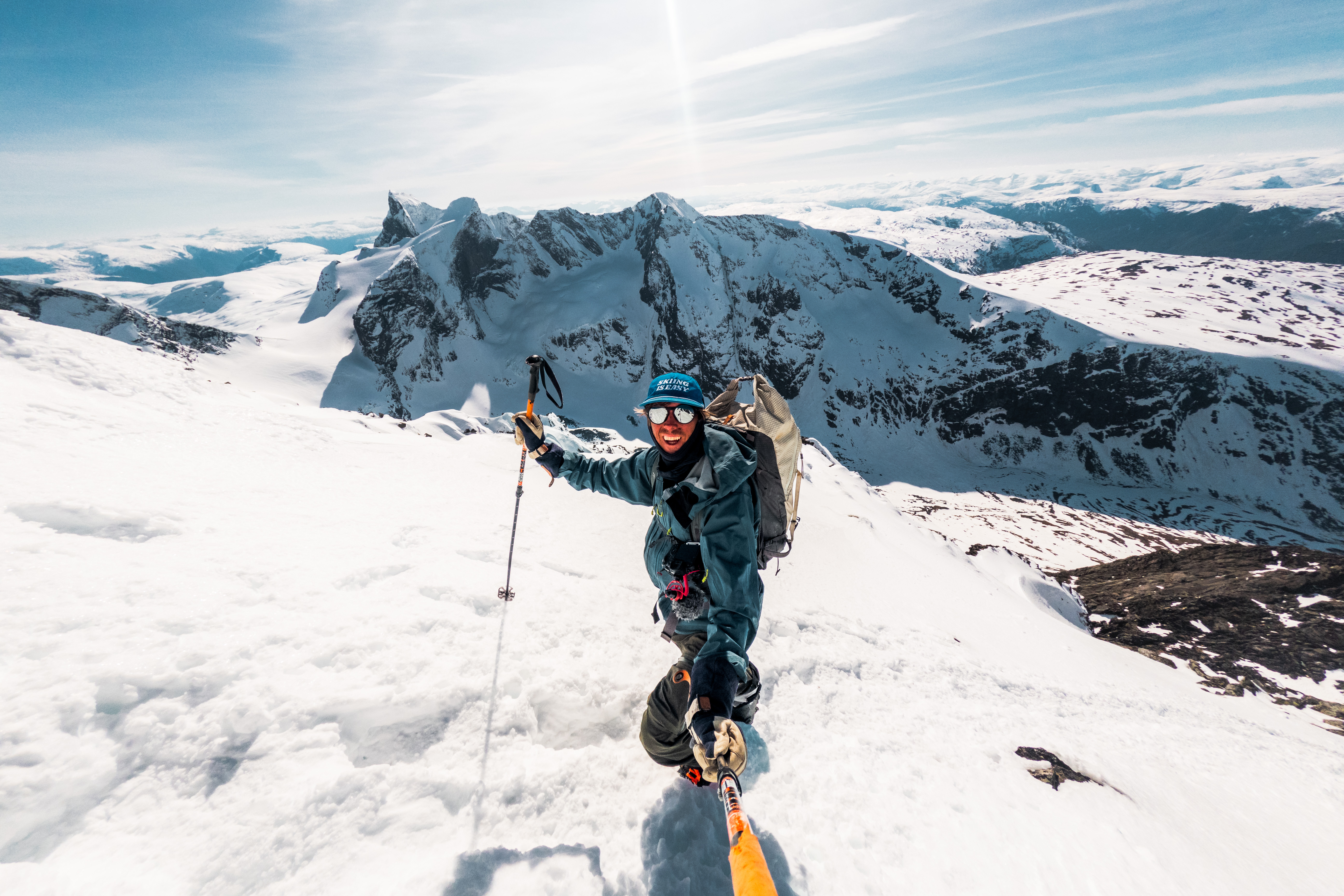 En glad mann med solbriller og caps foran snødekte fjell i bakgrunnen.