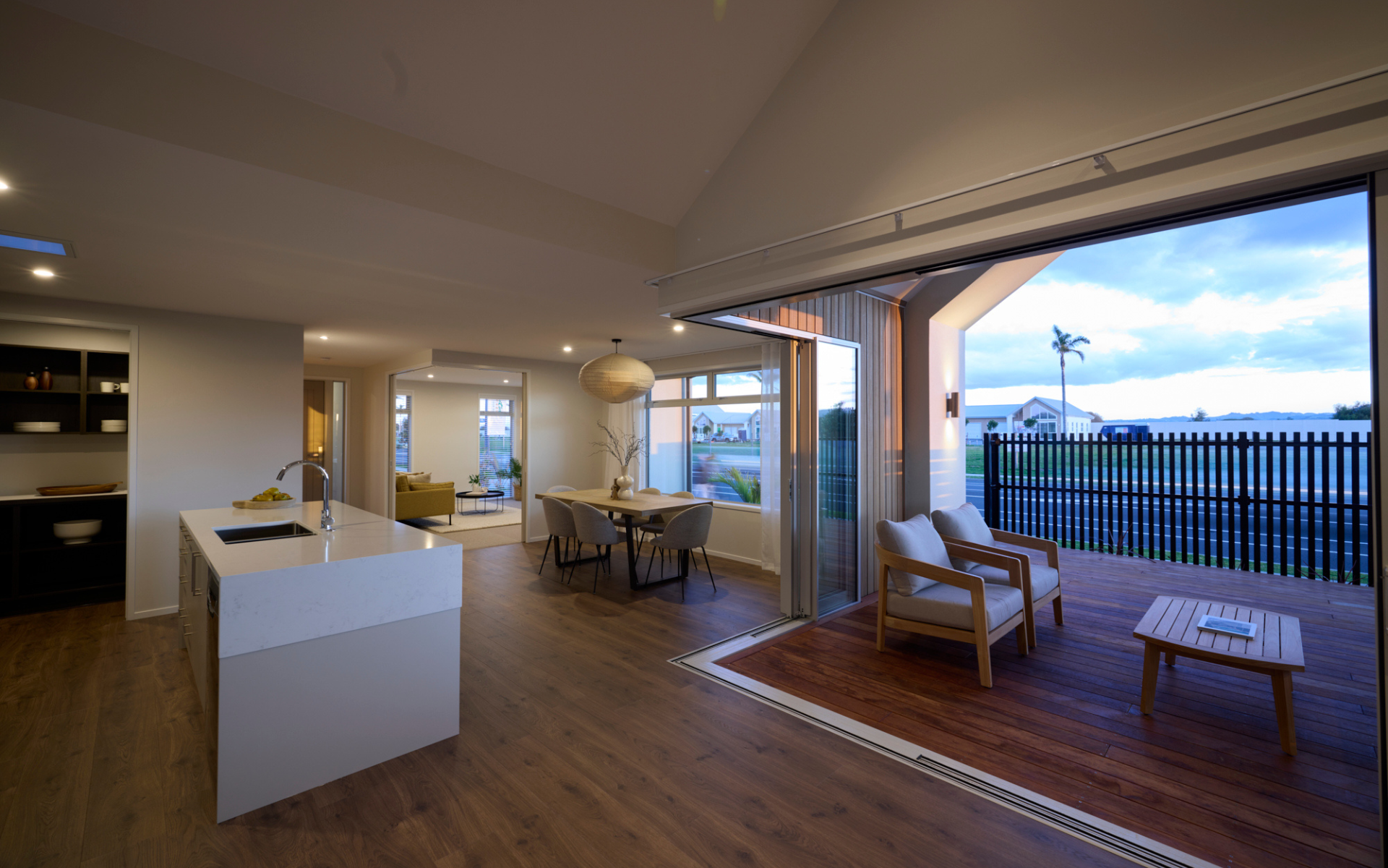 Contemporary kitchen and dining space with wood flooring, stone island, and large sliding doors opening to a timber deck with lounge chairs and outdoor views.