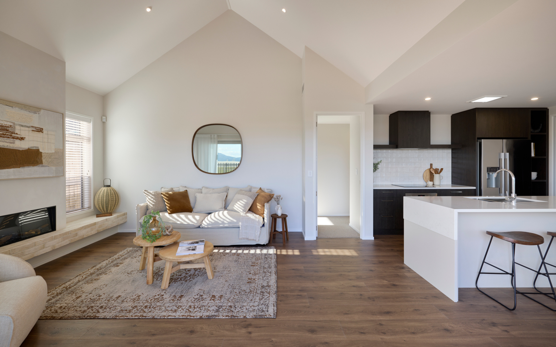 Contemporary living space with a neutral sofa, textured rug, and feature mirror, opening into a stylish kitchen with dark cabinetry and stone island bench