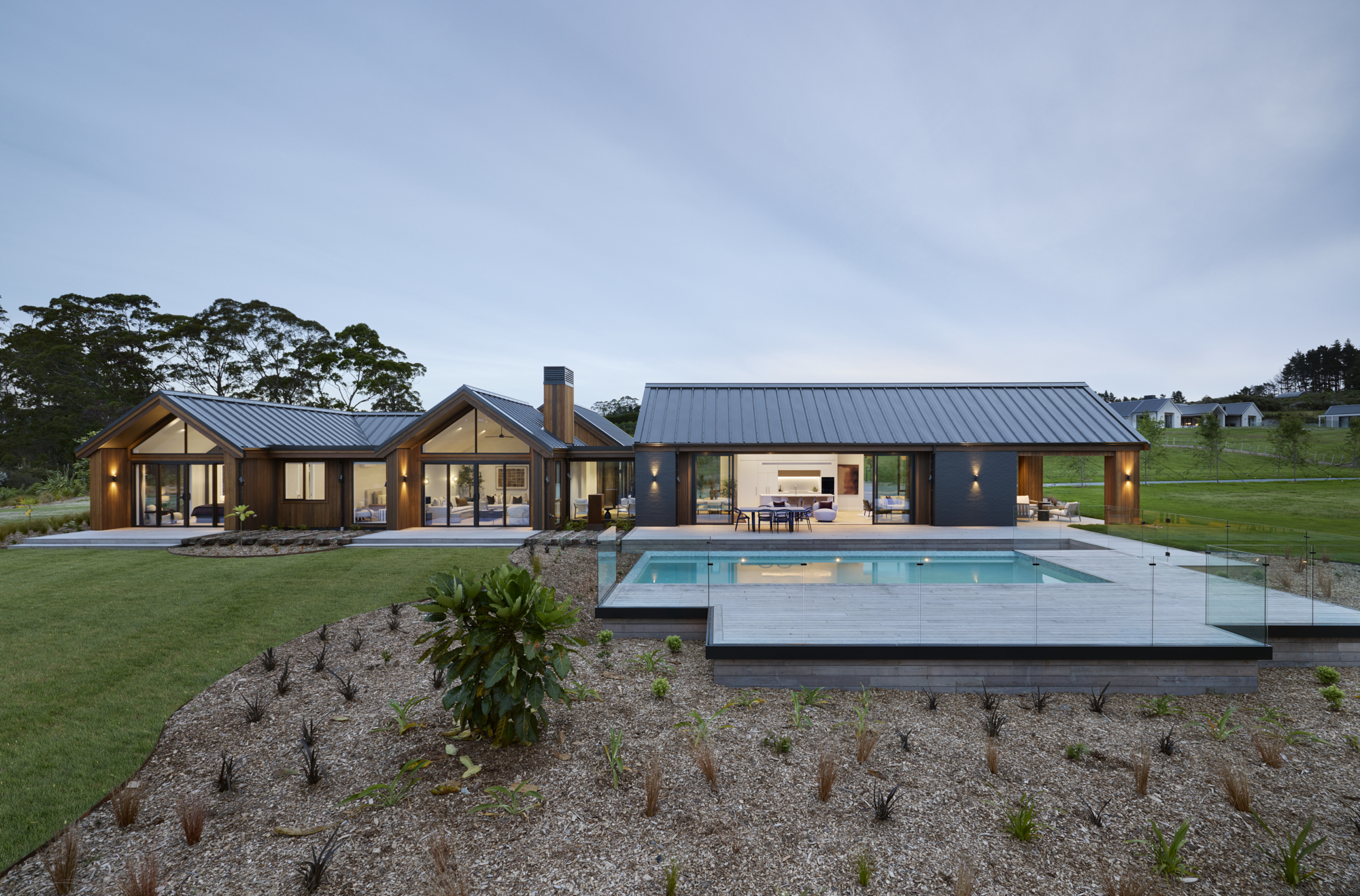 Modern architectural home with cedar cladding, dark rooflines, and a swimming pool overlooking landscaped gardens at dusk.