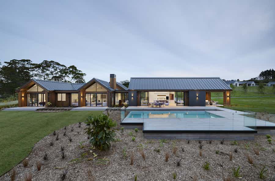 Modern architectural home with cedar cladding, dark rooflines, and a swimming pool overlooking landscaped gardens at dusk.