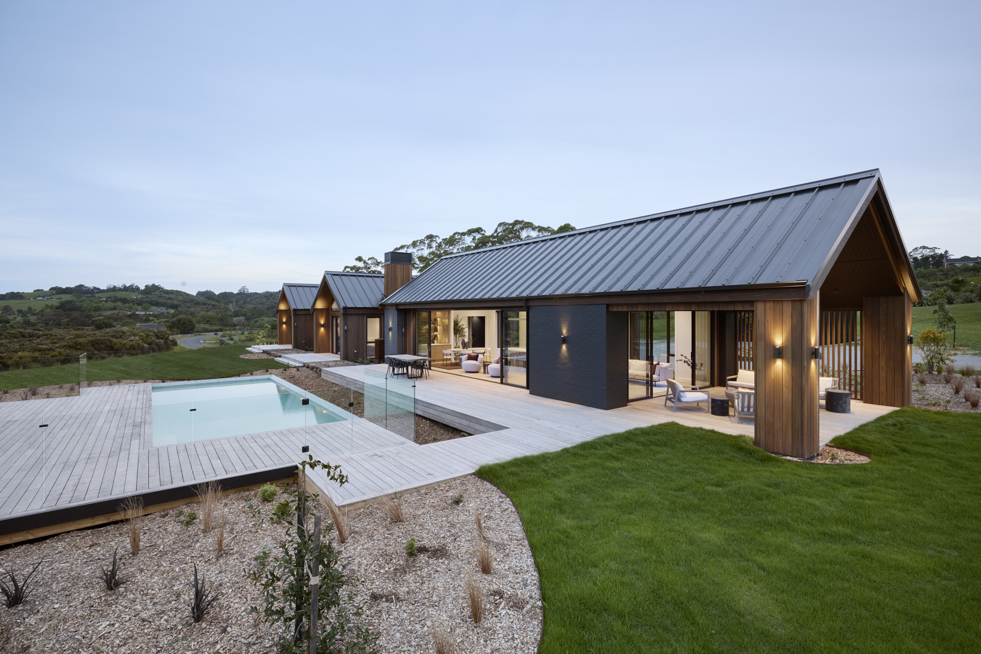 Contemporary cedar-clad home with expansive decking and a pool framed by glass balustrades in a rural New Zealand setting.