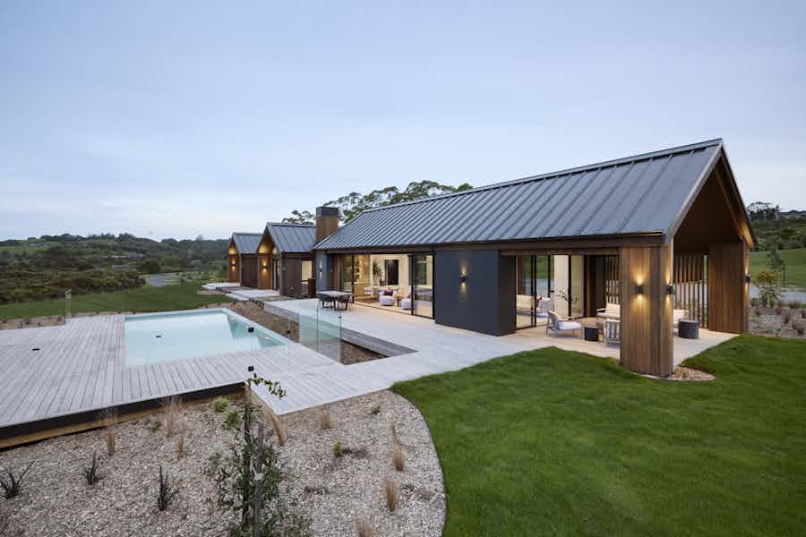 Contemporary cedar-clad home with expansive decking and a pool framed by glass balustrades in a rural New Zealand setting.
