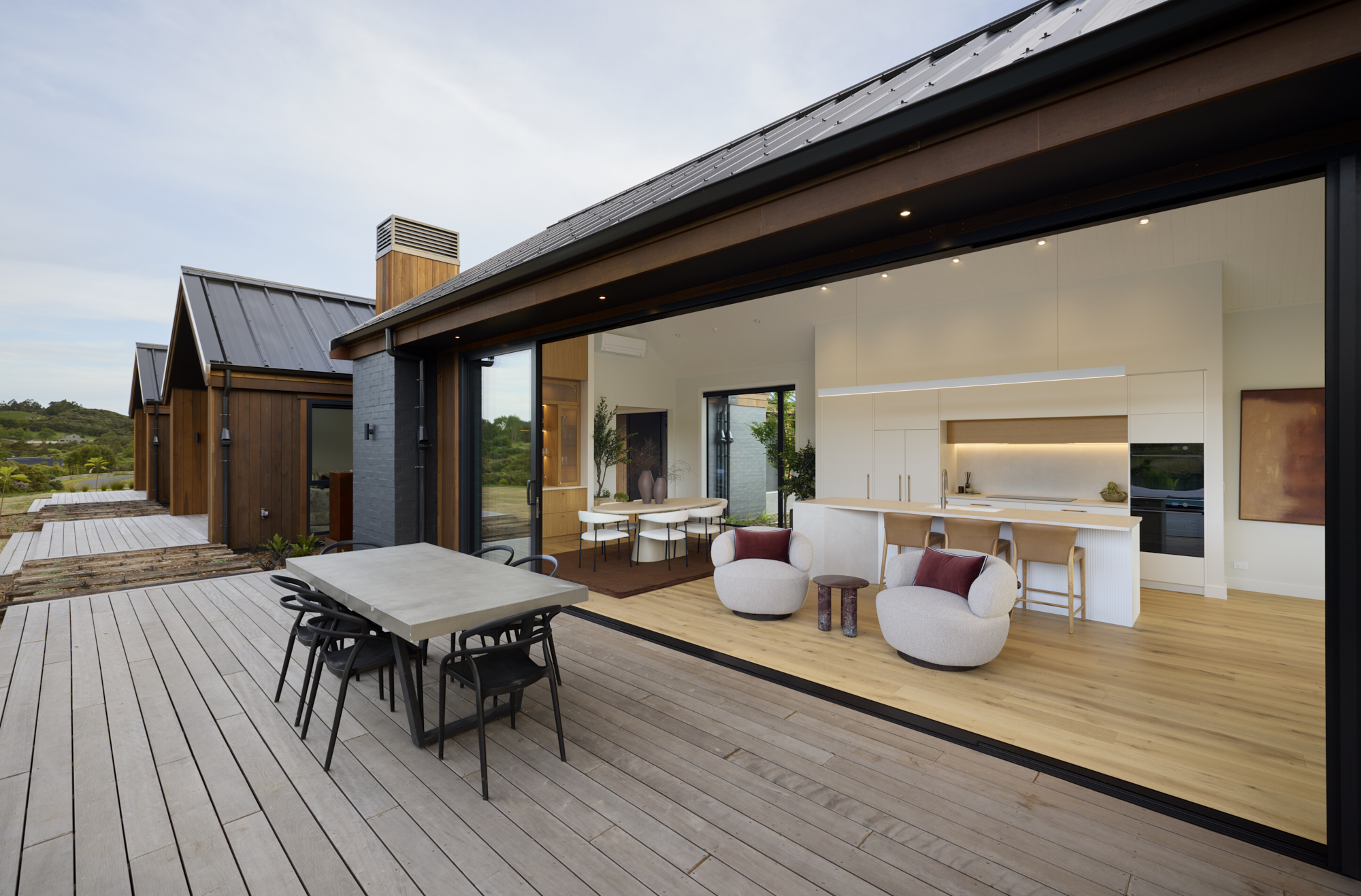 Outdoor dining area connected to an open-plan kitchen and living space with sliding glass doors and timber decking.