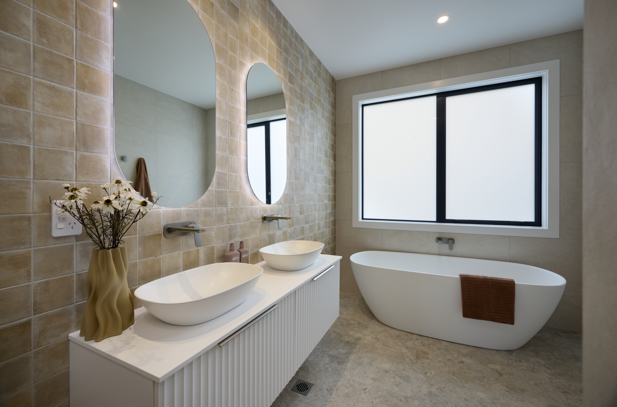 Modern bathroom with double vessel sinks, curved backlit mirrors, textured tile feature wall, and a freestanding bathtub beneath a large frosted window.