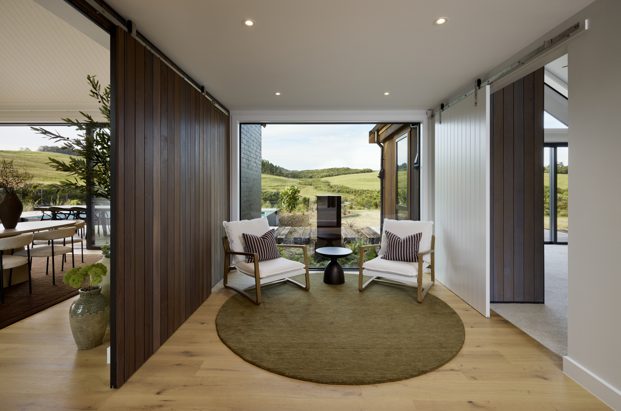 Cozy sitting nook with sliding timber feature panels, two armchairs, round rug, and floor-to-ceiling window framing rural landscape views
