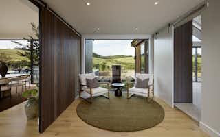 Cozy sitting nook with sliding timber feature panels, two armchairs, round rug, and floor-to-ceiling window framing rural landscape views