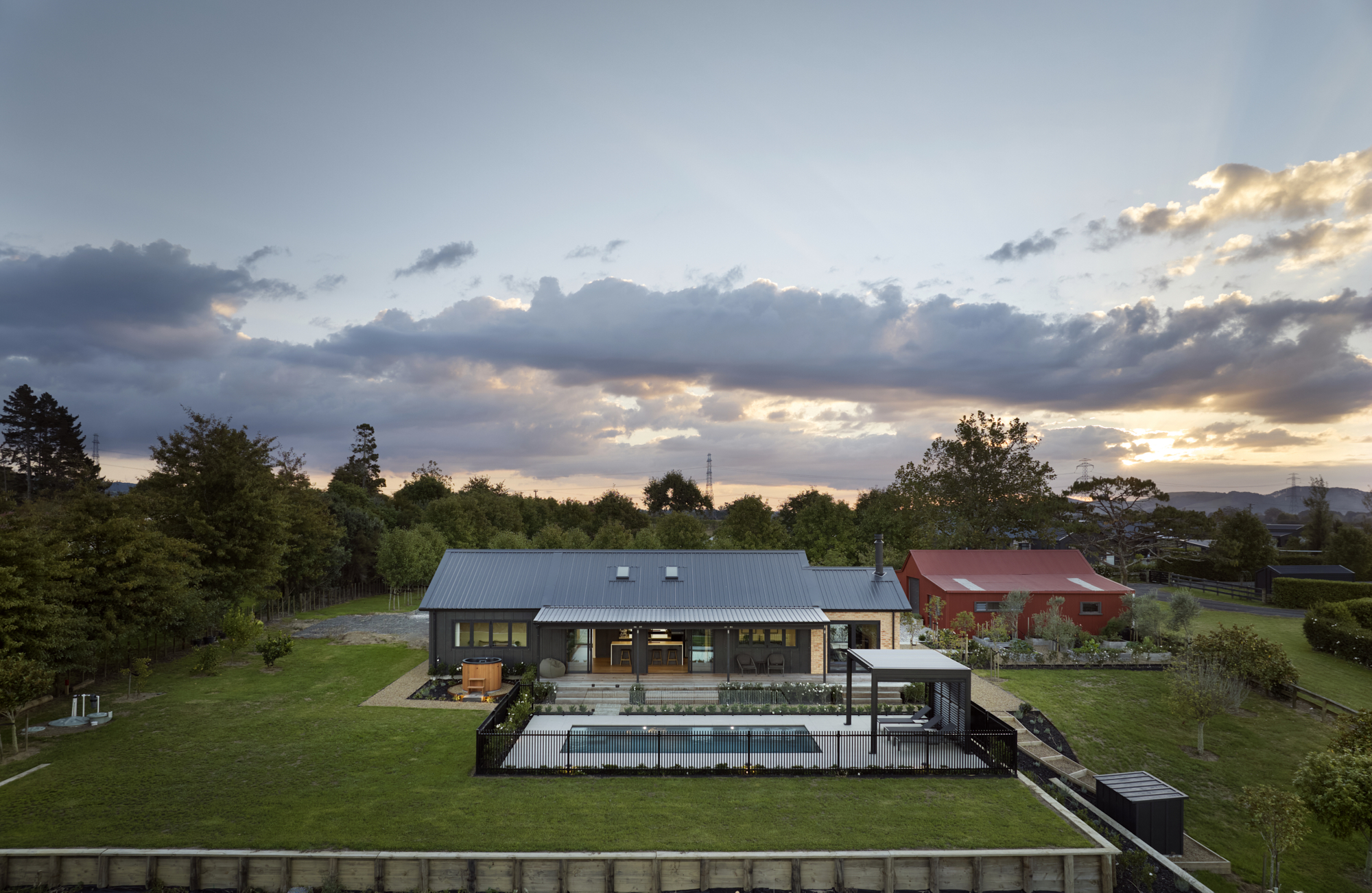 Aerial dusk view of the Signature Homes Clevedon House Plan, showing a dark-roofed single-storey home with pool, hot tub, and lush gardens against a dramatic sunset sky.