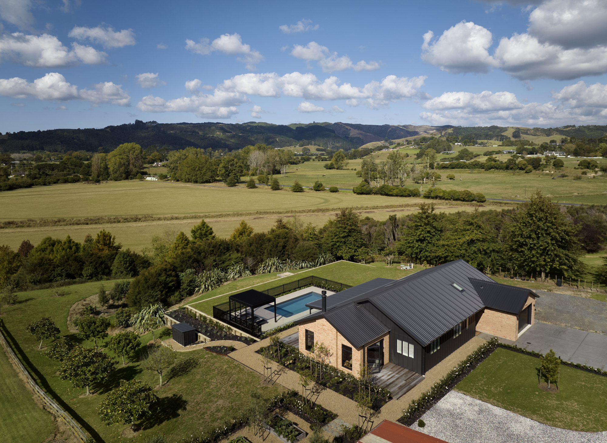 Aerial daytime view of the Clevedon home surrounded by rolling green New Zealand farmland, with a pool, pergola, and established gardens visible from above.