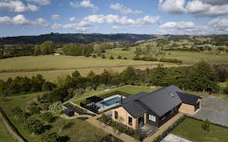 Aerial daytime view of the Clevedon home surrounded by rolling green New Zealand farmland, with a pool, pergola, and established gardens visible from above.