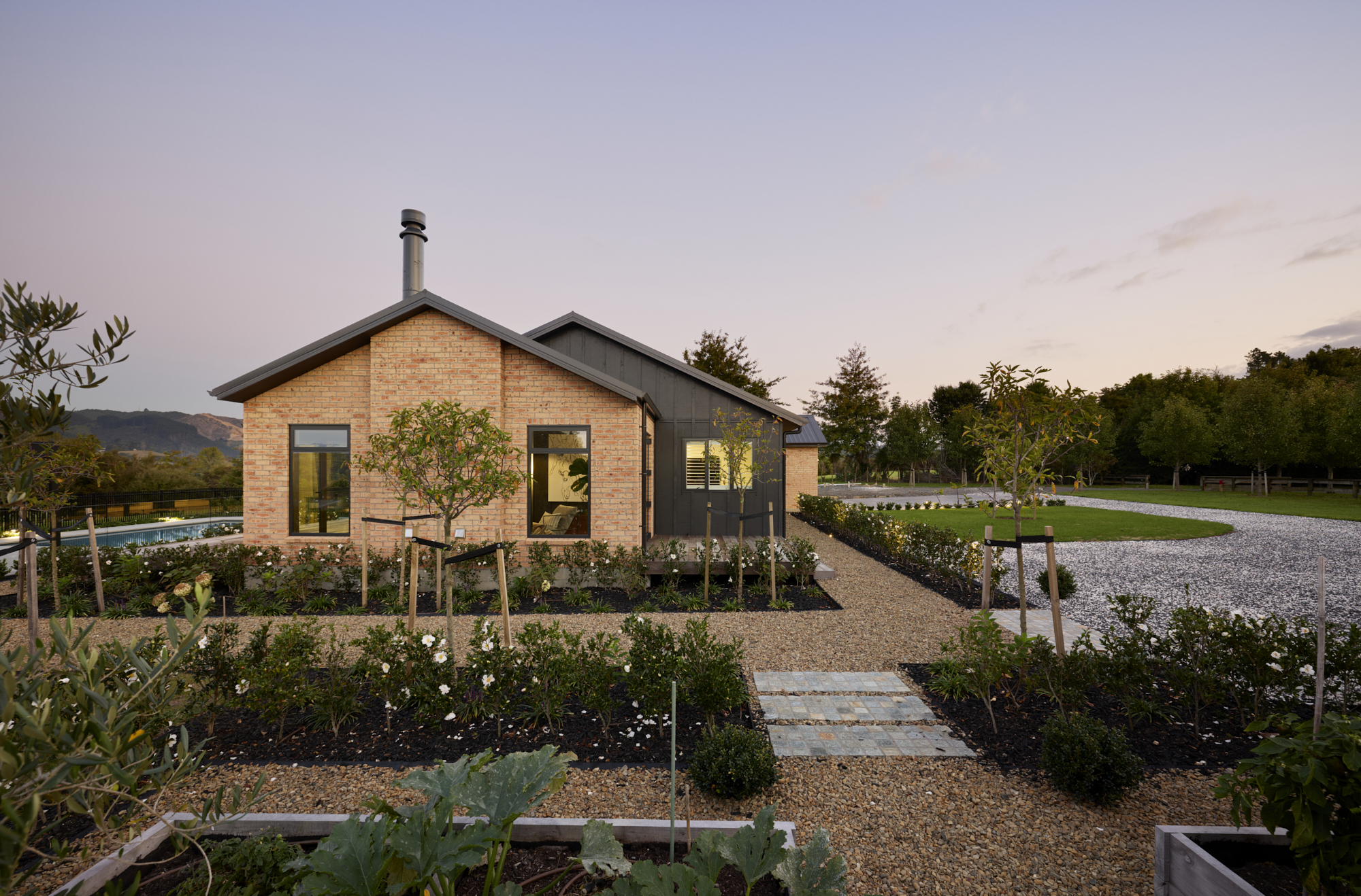 Ground-level view of the brick and dark cladding gable end of the Clevedon home, framed by raised vegetable garden beds, gravel paths, and a pool to the left.