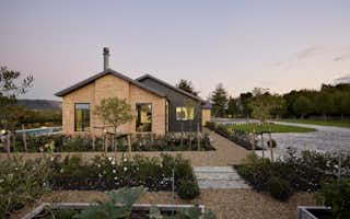 Ground-level view of the brick and dark cladding gable end of the Clevedon home, framed by raised vegetable garden beds, gravel paths, and a pool to the left.