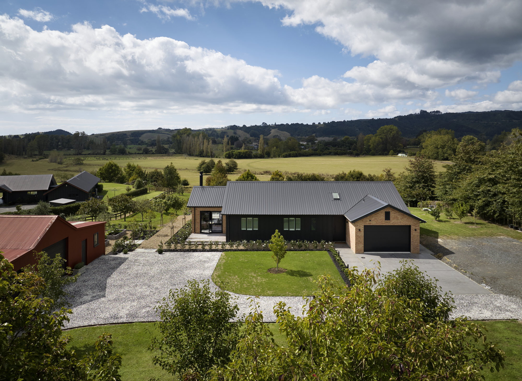 Elevated view of the Clevedon home's front aspect showing brick and dark board-and-batten exterior, double garage, gravel driveway, and wide rural valley views beyond.