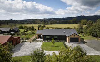 Elevated view of the Clevedon home's front aspect showing brick and dark board-and-batten exterior, double garage, gravel driveway, and wide rural valley views beyond.