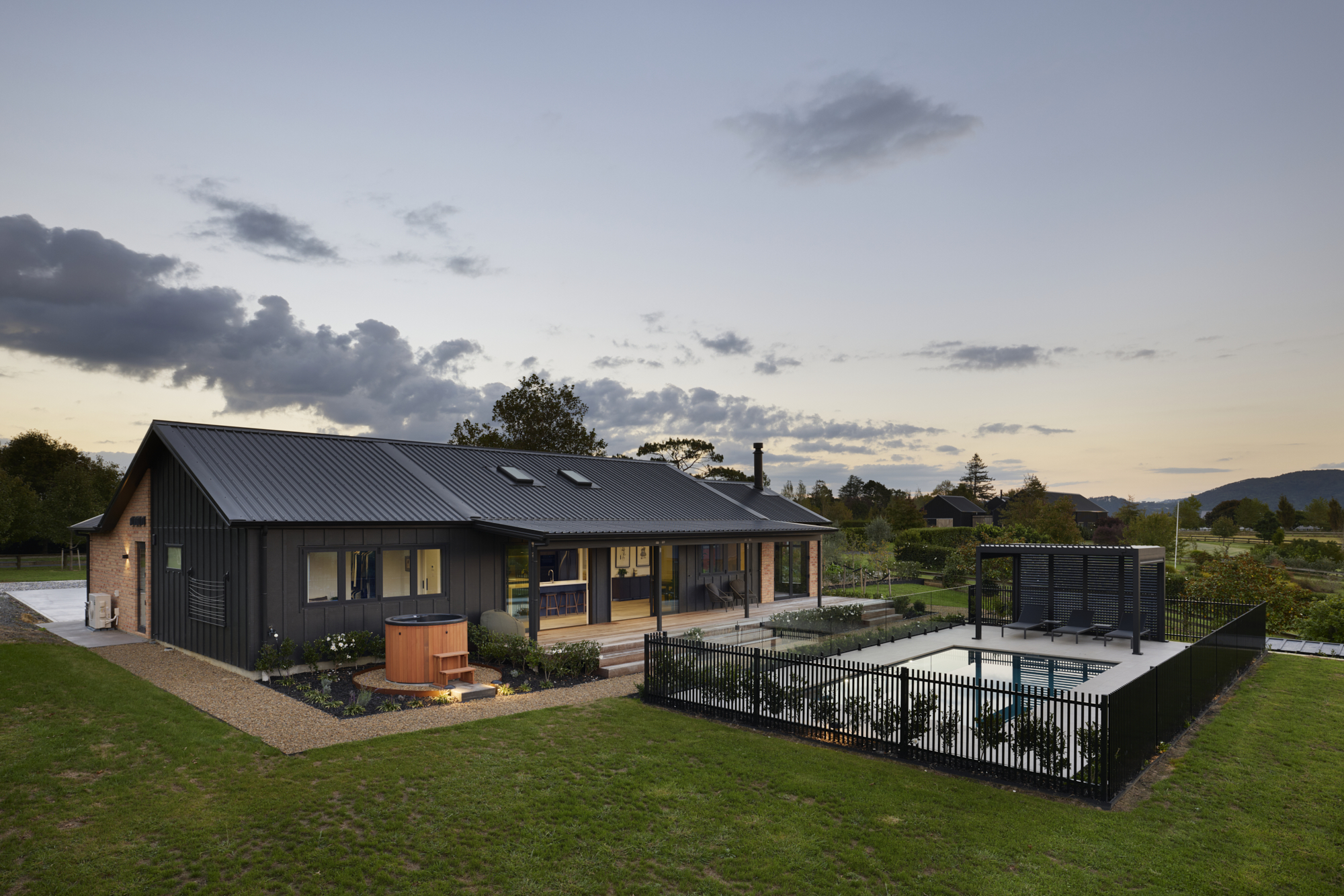 Dusk front facade of the Clevedon home showing the full length of the dark board-and-batten exterior with warm interior lighting, gravel driveway, and white flowering garden border.