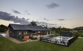 Dusk front facade of the Clevedon home showing the full length of the dark board-and-batten exterior with warm interior lighting, gravel driveway, and white flowering garden border.