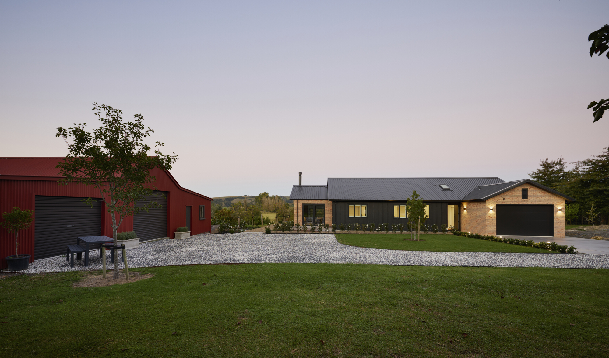 Dusk view from across the yard showing the Clevedon home alongside a red-roofed farm shed, with wide open lawns and a soft purple evening sky.
