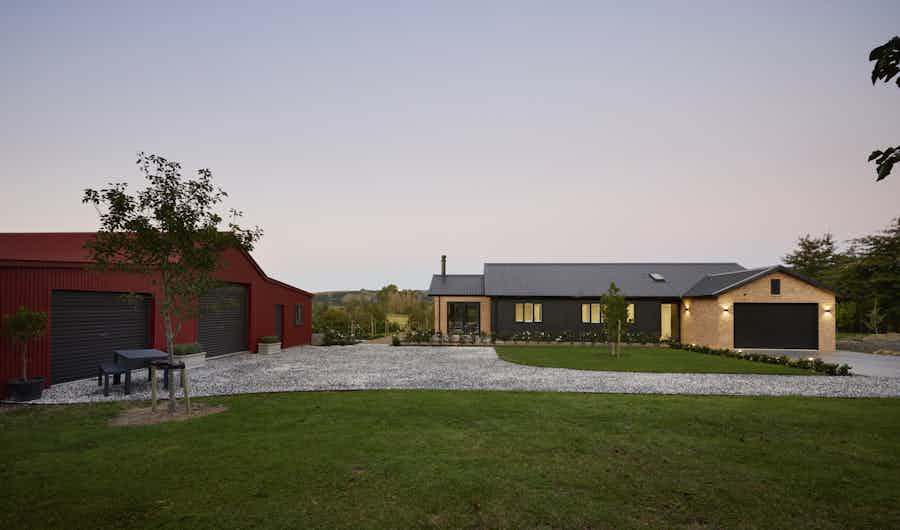 Dusk view from across the yard showing the Clevedon home alongside a red-roofed farm shed, with wide open lawns and a soft purple evening sky.