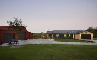 Dusk view from across the yard showing the Clevedon home alongside a red-roofed farm shed, with wide open lawns and a soft purple evening sky.