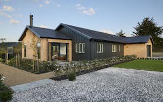 Daytime front facade of the Clevedon home featuring warm brick gable ends, dark cladding, plantation shutter windows, a welcoming entry deck, and gravel forecourt.