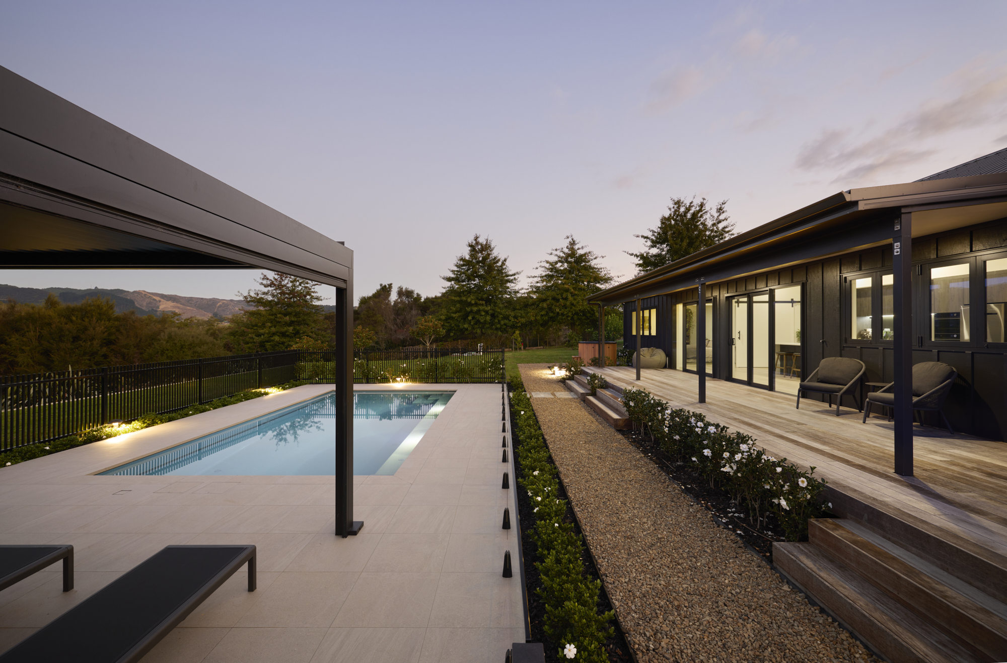 Dusk view of the pool and outdoor entertaining area, with the Clevedon home's open-plan living interior visible through large sliding glass doors and a pergola to the left.