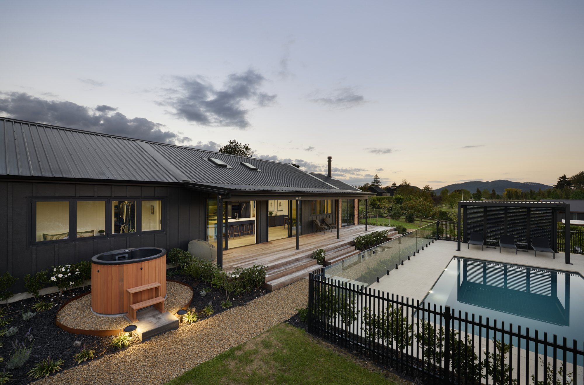 Dusk rear view of the Clevedon home showing the cedar hot tub, outdoor deck, pool with black fencing, and open-plan living areas bathed in warm interior light.