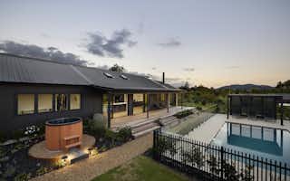 Dusk rear view of the Clevedon home showing the cedar hot tub, outdoor deck, pool with black fencing, and open-plan living areas bathed in warm interior light.