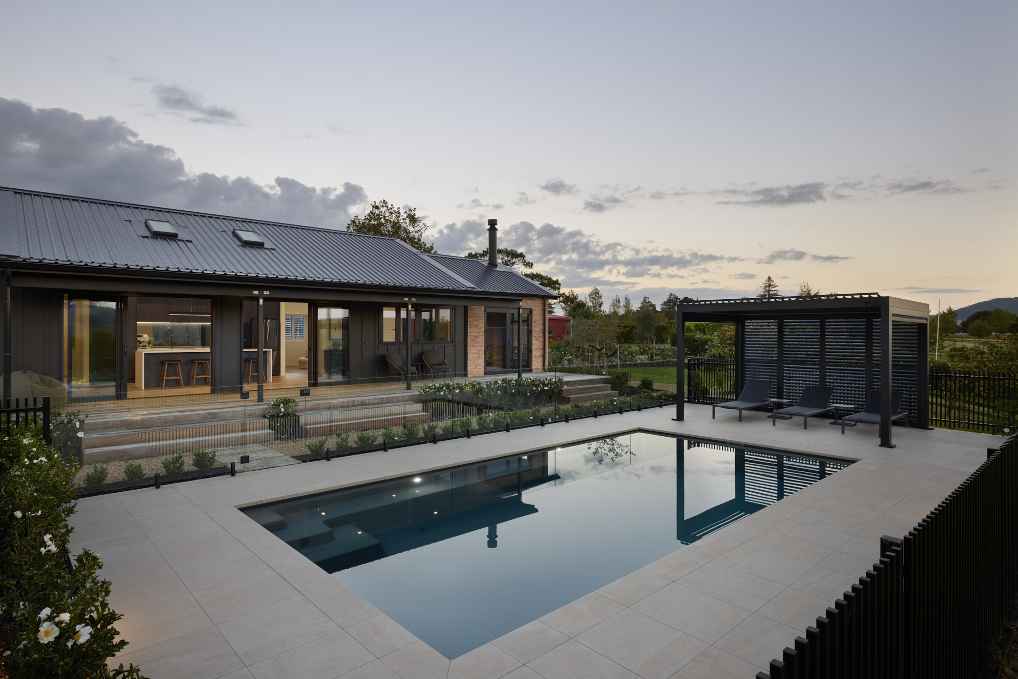 Dusk pool terrace view of the Clevedon home, with still water reflecting the sky, sun loungers under a louvred pergola, and open-plan interiors visible beyond.