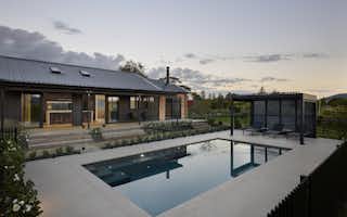 Dusk pool terrace view of the Clevedon home, with still water reflecting the sky, sun loungers under a louvred pergola, and open-plan interiors visible beyond.