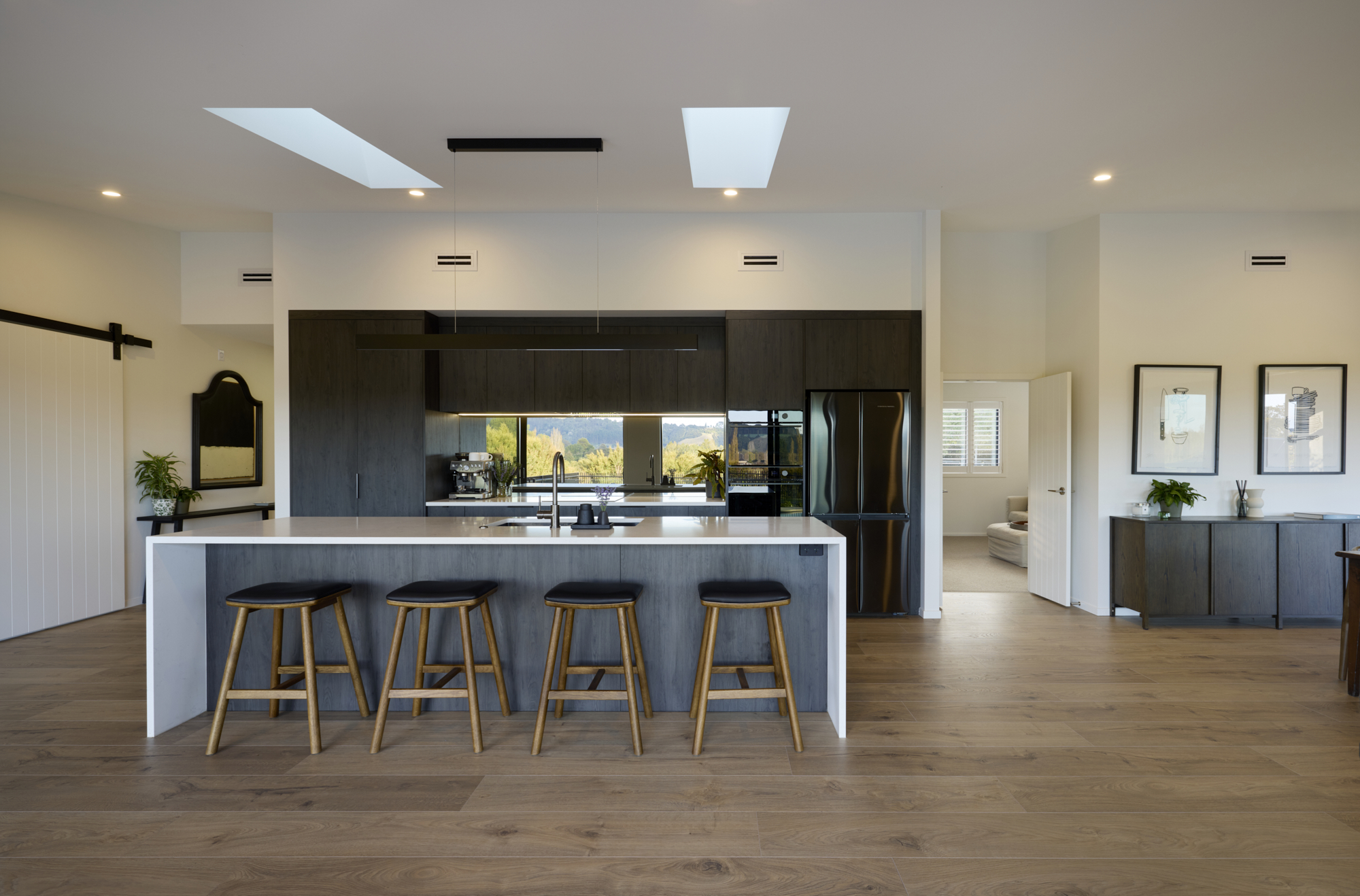 Wide interior shot of the designer kitchen with dark cabinetry, stone island bench, skylight, and open-plan flow through to the dining area and brick fireplace beyond.