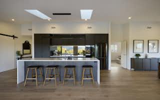 Wide interior shot of the designer kitchen with dark cabinetry, stone island bench, skylight, and open-plan flow through to the dining area and brick fireplace beyond.