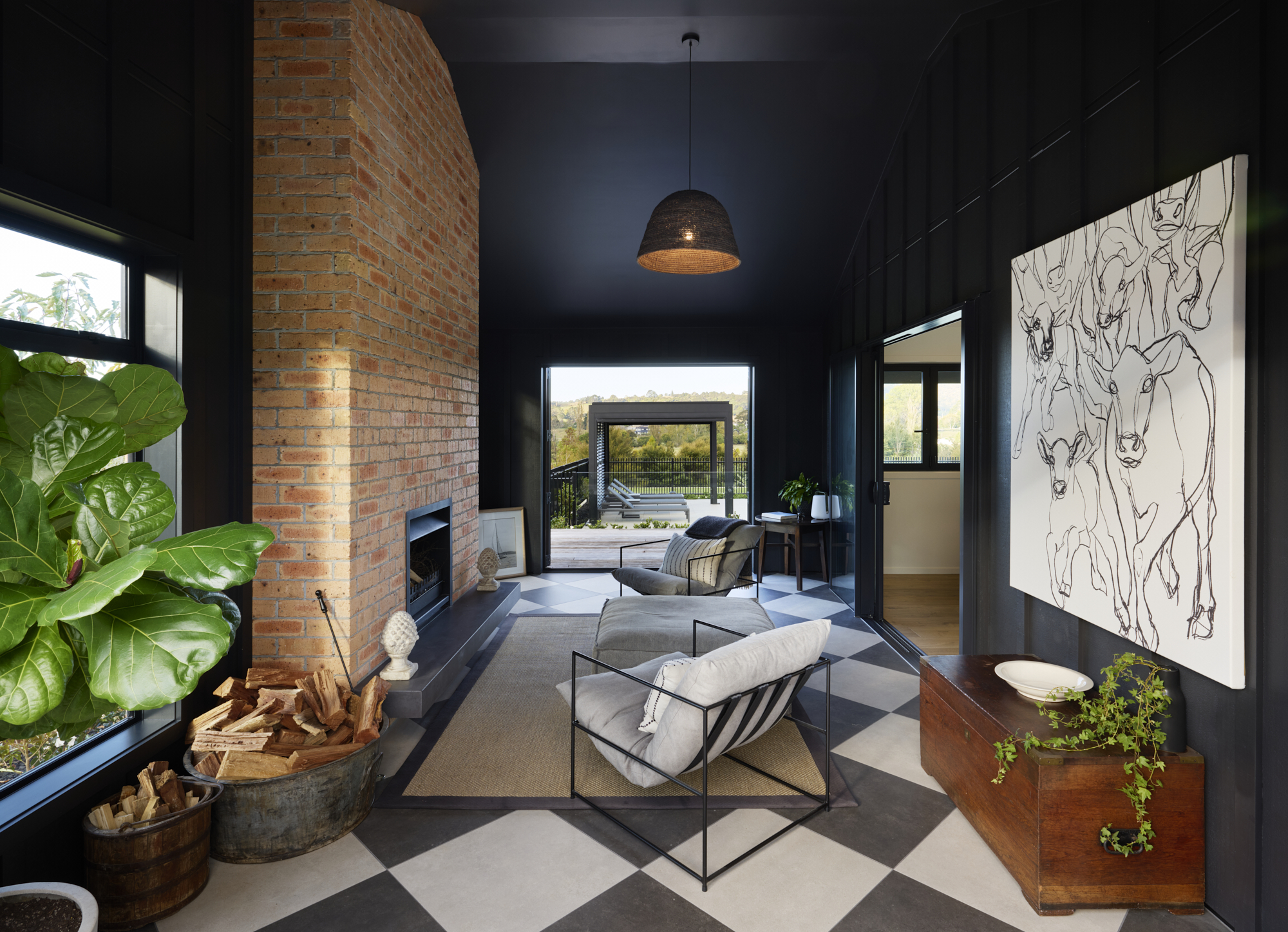 Dramatic dark-painted sunken lounge with exposed brick fireplace, checkerboard concrete floor, woven pendant light, large-scale cattle artwork, and open bifold doors to the outdoor deck.