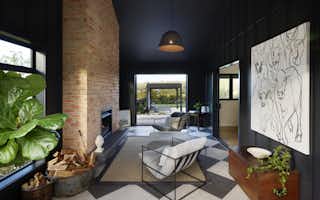 Dramatic dark-painted sunken lounge with exposed brick fireplace, checkerboard concrete floor, woven pendant light, large-scale cattle artwork, and open bifold doors to the outdoor deck.